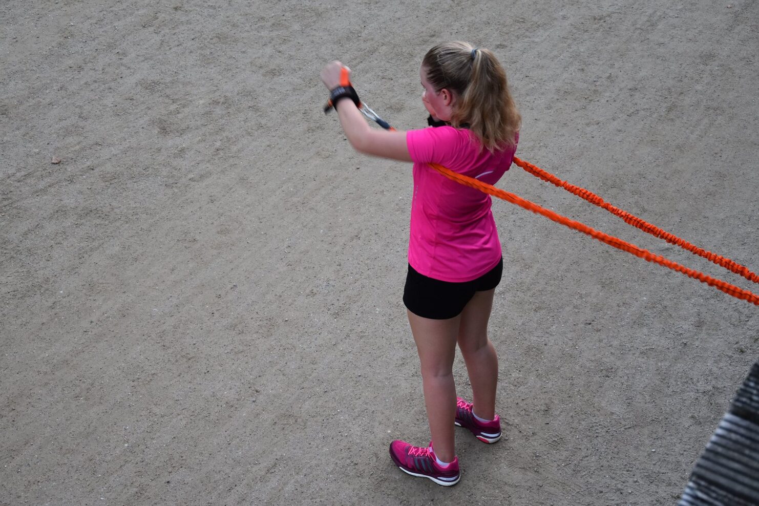 Sportlerin in pinkem Shirt und schwarzen Shorts zieht orangefarbene Widerstandsb&auml;nder auf einem sandigen Untergrund.