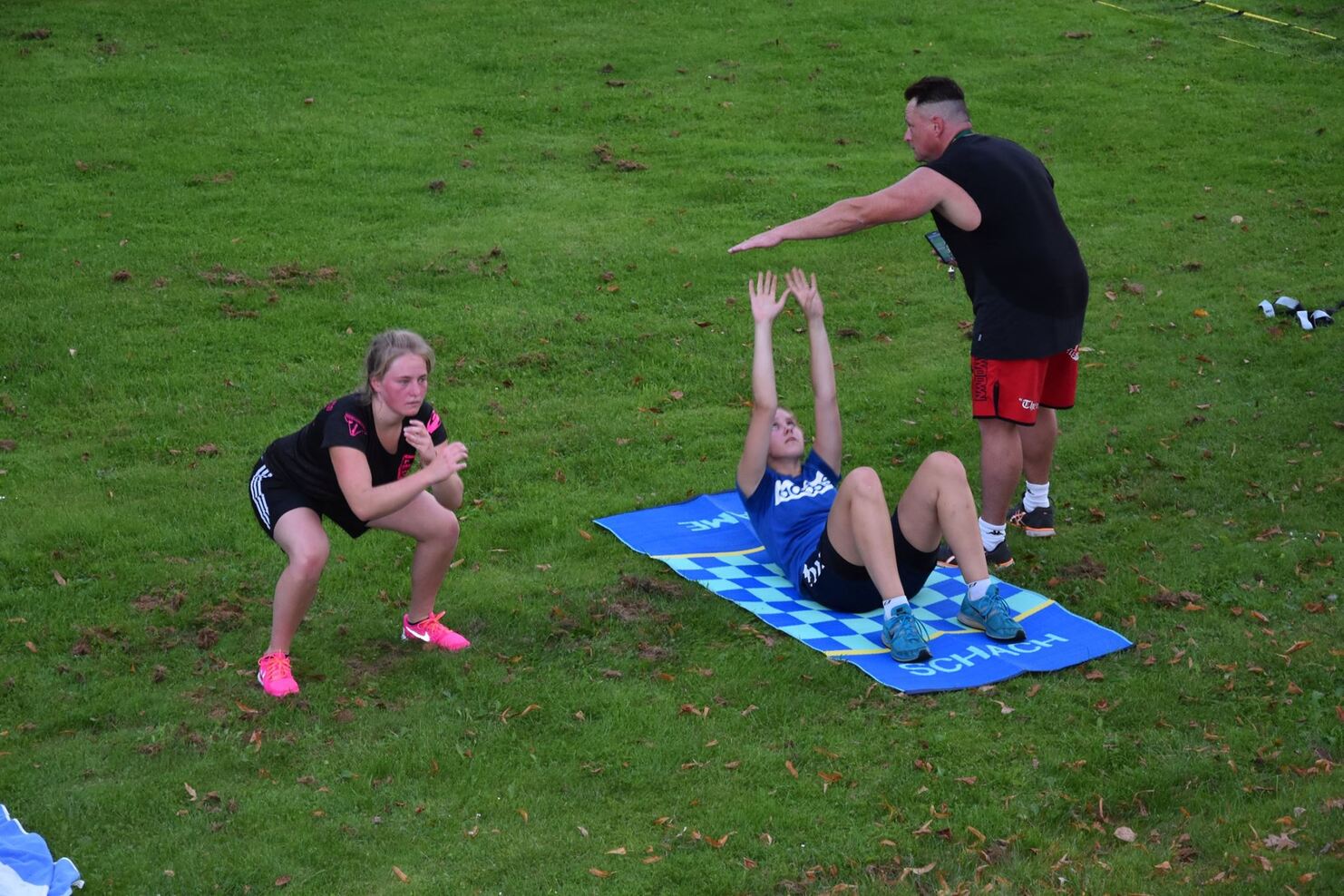 Drei Personen trainieren auf einem gr&uuml;nen Rasen: zwei machen &Uuml;bungen, w&auml;hrend ein Trainer Anweisungen gibt.