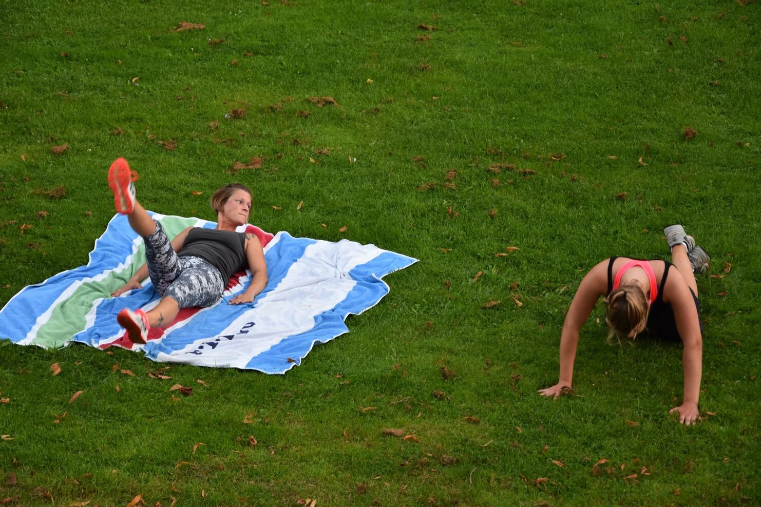 Zwei Frauen trainieren im Freien: Eine macht Bein&uuml;bungen auf einer Decke, die andere f&uuml;hrt Liegest&uuml;tze aus.
