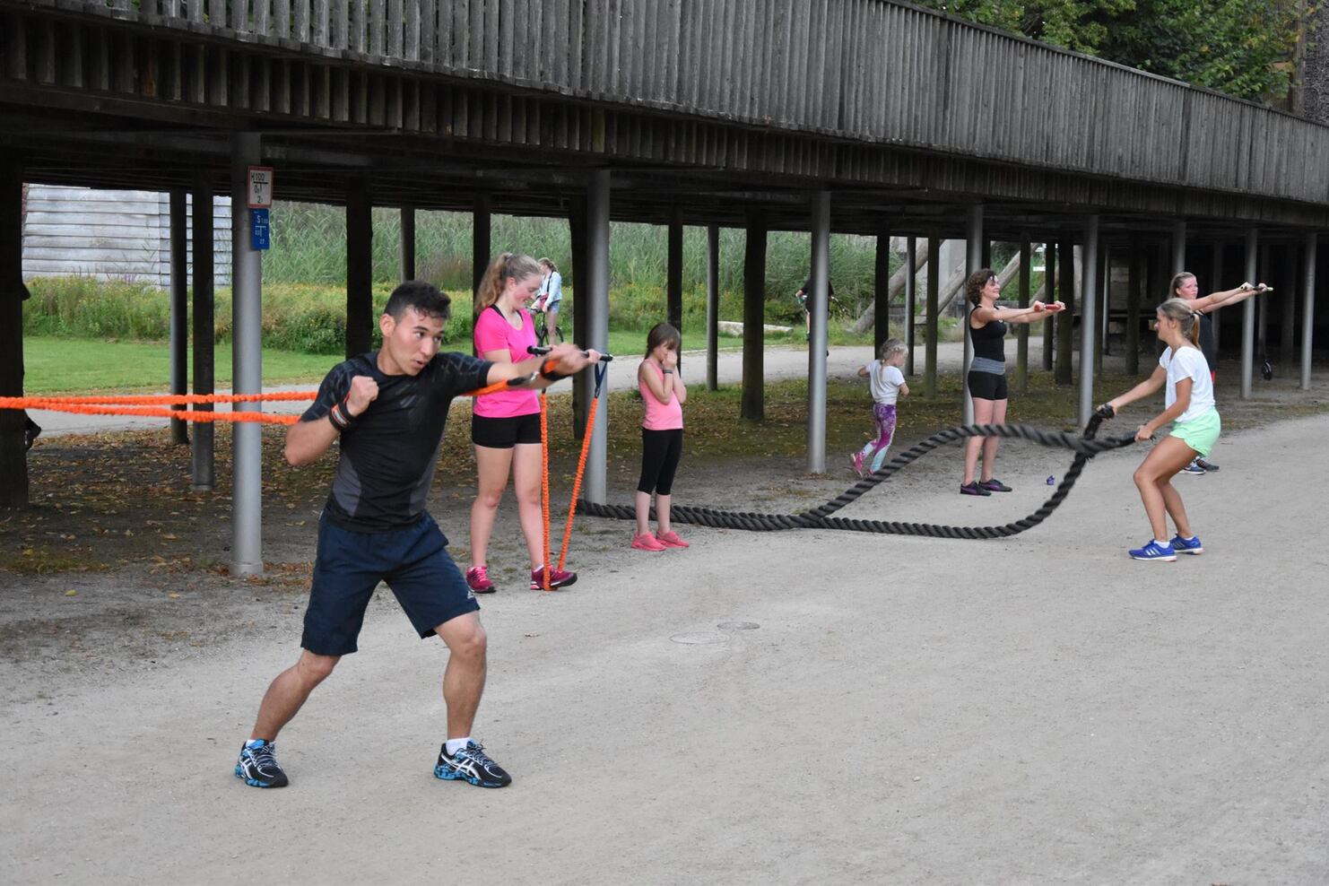 Gruppe von Kindern und einem Erwachsenen trainiert mit Widerstandsb&auml;ndern und einem Schlauch auf einem Sportplatz im Freien.