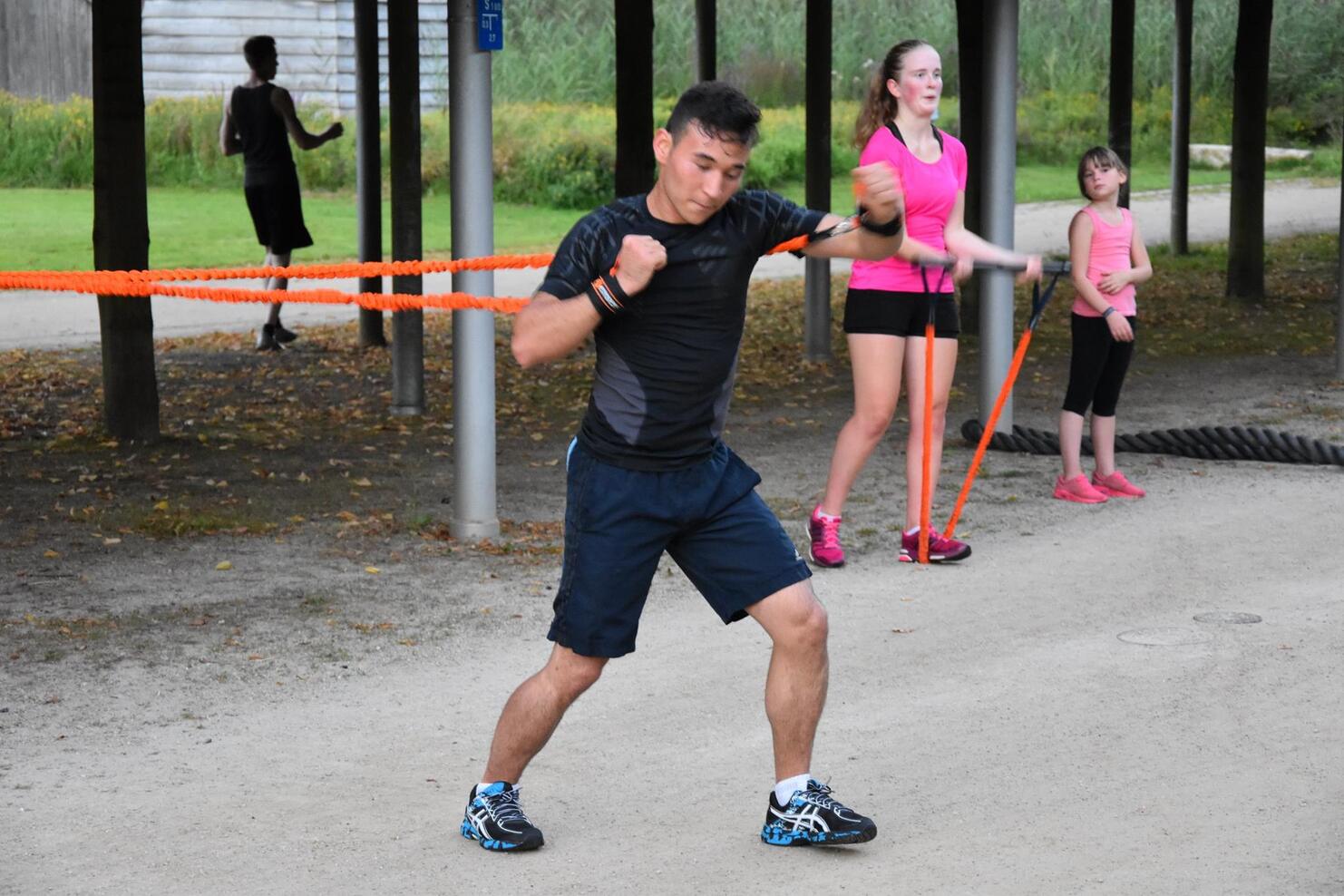 Mann trainiert mit Widerstandsb&auml;ndern im Freien, umgeben von zwei Frauen und einem M&auml;dchen, die ebenfalls aktiv sind.