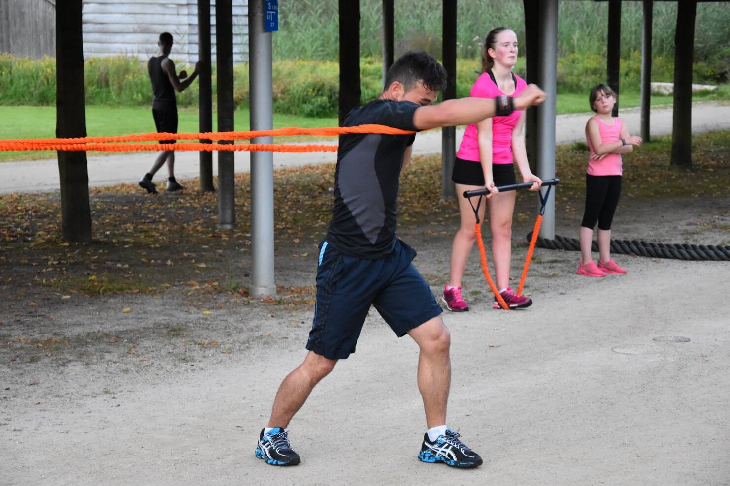 Gruppentraining im Freien: Ein Mann dehnt mit Widerstandsband, w&auml;hrend zwei Frauen und ein M&auml;dchen bereitstehen.