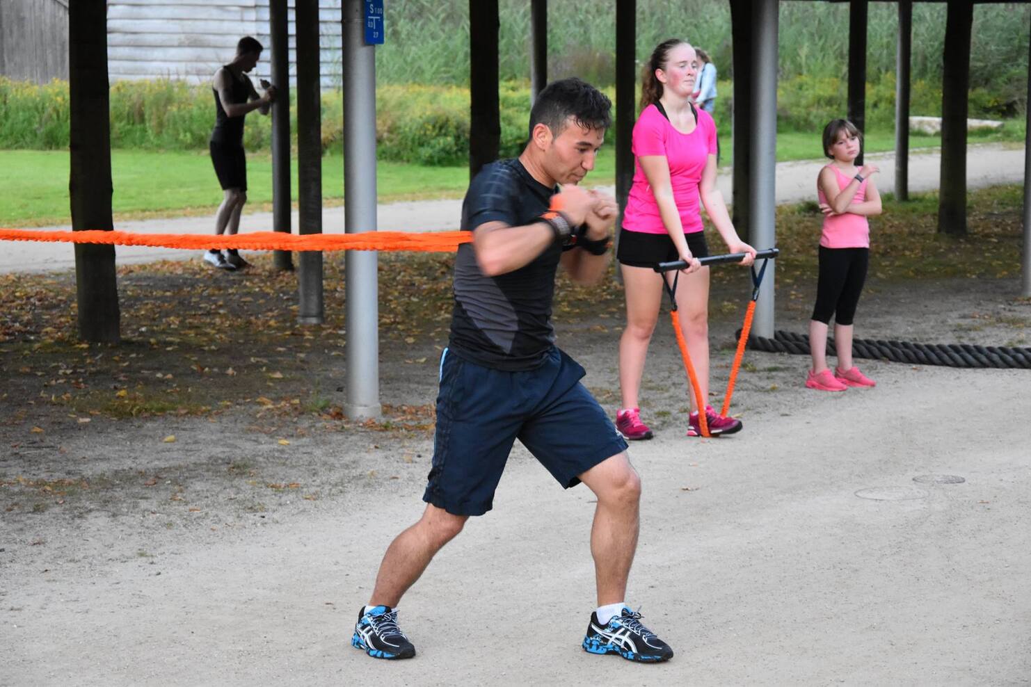 Mann trainiert mit Widerstandsb&auml;ndern im Freien, w&auml;hrend Frauen in Sportkleidung zuschauen und ebenfalls trainieren.
