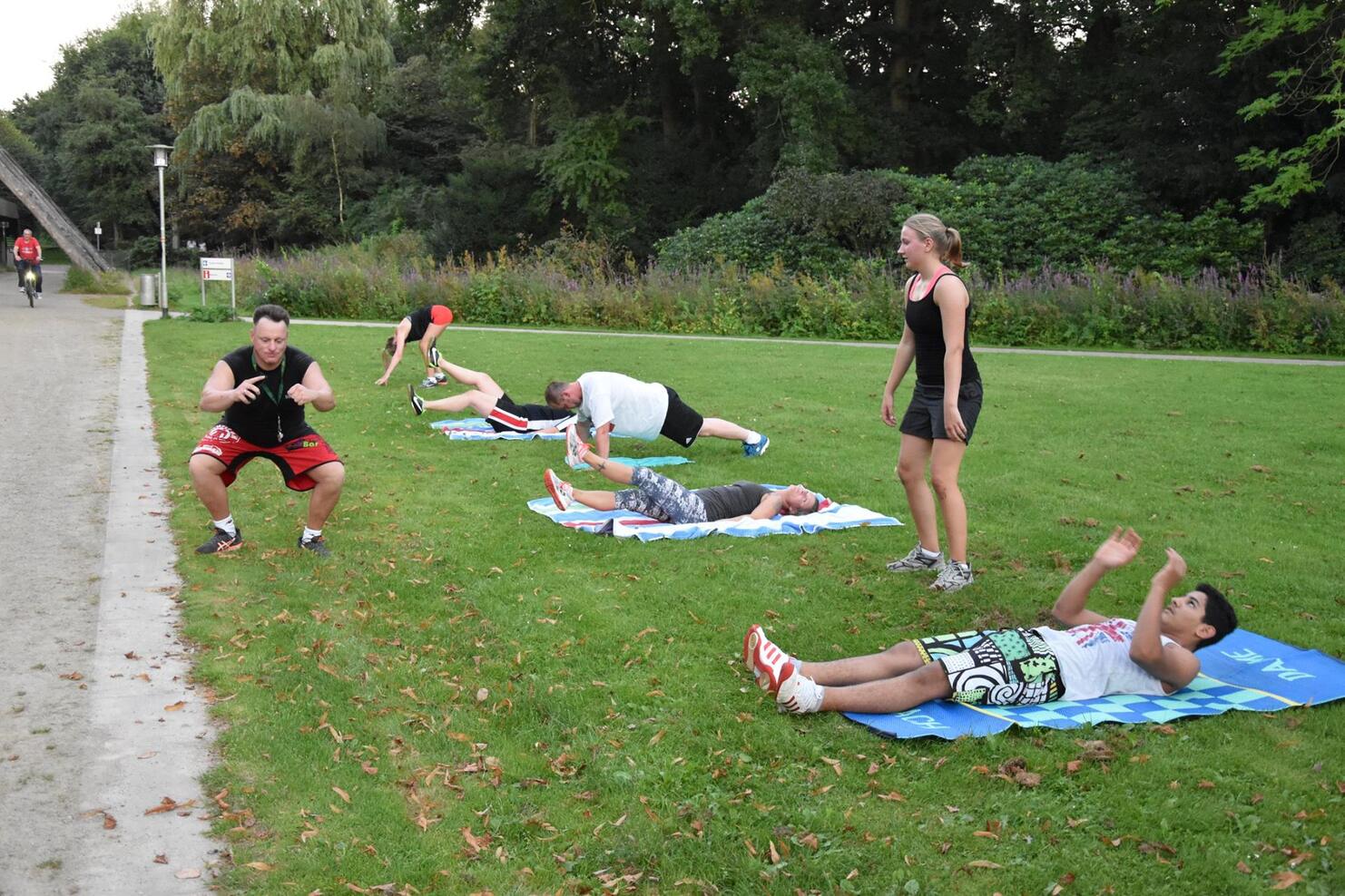 Gruppe von sechs Personen, die auf gr&uuml;ner Wiese verschiedene Fitness&uuml;bungen machen, umgeben von B&auml;umen.