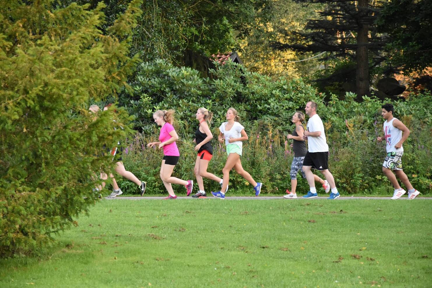 Gruppe von acht Menschen joggt auf einem Weg in einem park&auml;hnlichen Umfeld mit B&auml;umen im Hintergrund.