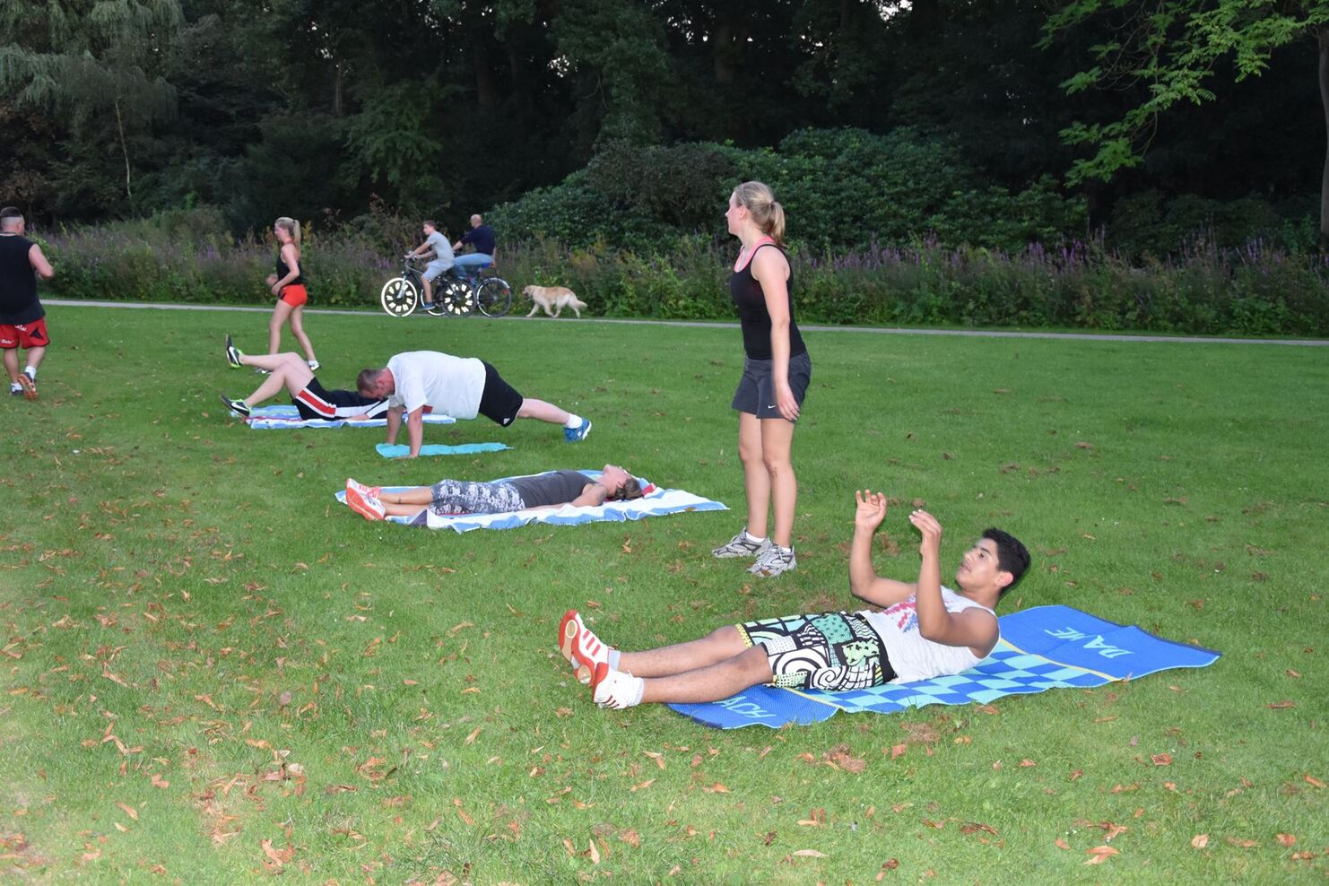 Mehrere Personen trainieren im Freien auf Matten in einem Park, einige machen Liegest&uuml;tze, andere &Uuml;bungen im Liegen.