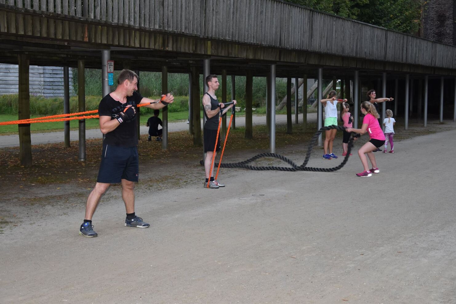 Gruppe von f&uuml;nf Personen trainiert im Freien mit Widerstandsb&auml;ndern und einem schweren Seil auf einem sandigen Weg.