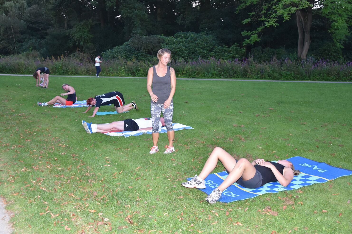 Gruppentraining im Park mit mehreren Personen auf Sportmatten, eine Trainerin beobachtet.