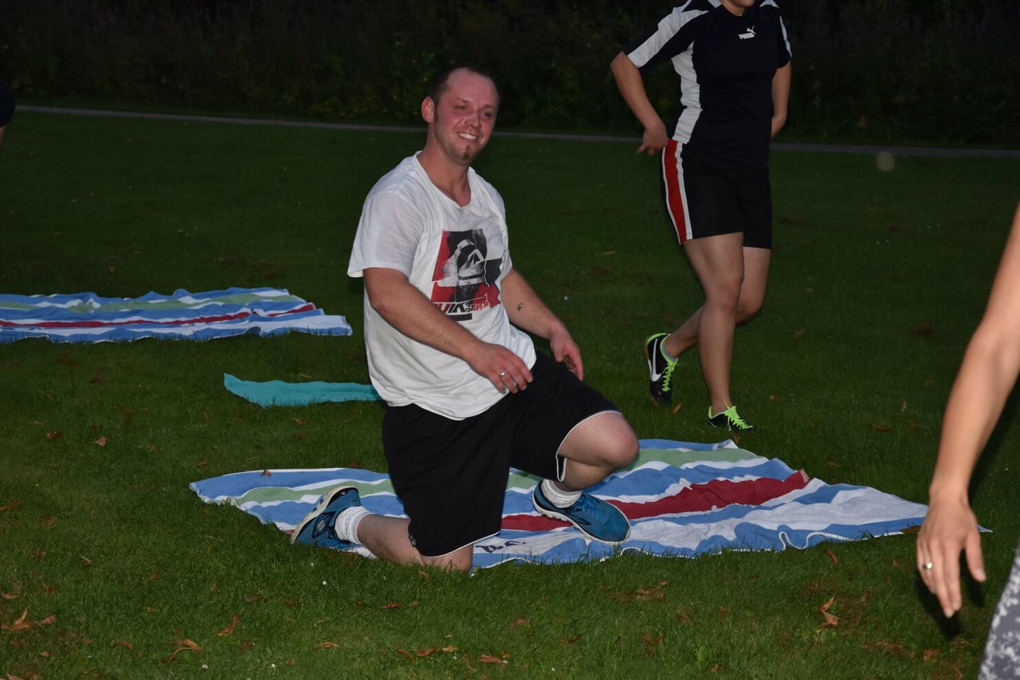 Mann kniet auf einer Wiese, tr&auml;gt ein T-Shirt und kurze Hose, w&auml;hrend eine Person im Hintergrund aktiv ist.