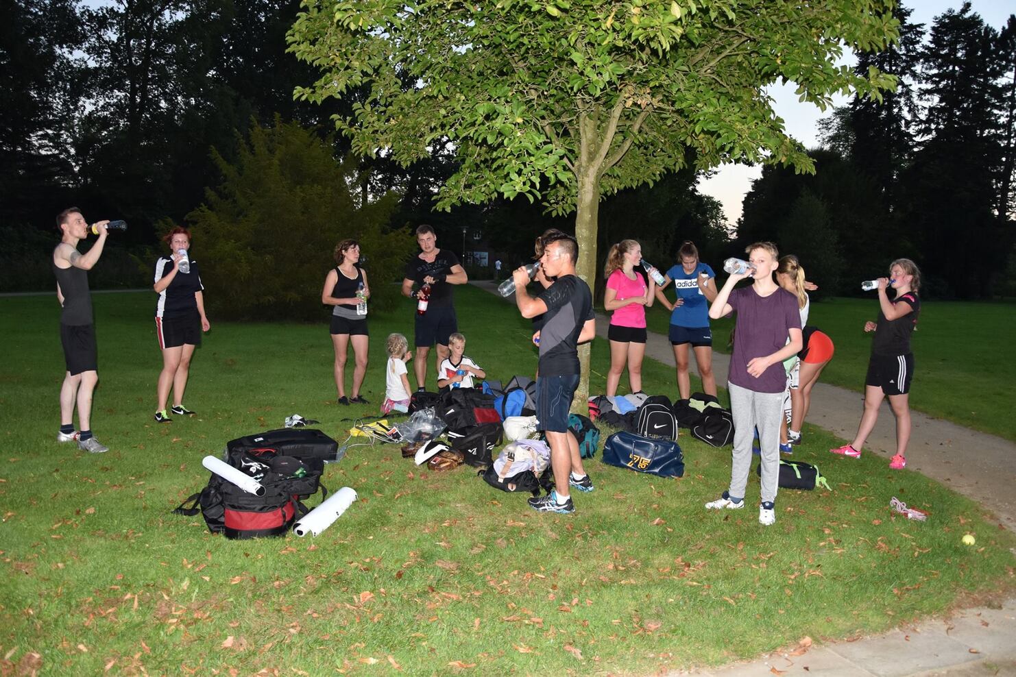 Gruppe von Sportlern in Sportkleidung trinkt Wasser und steht in einem Park unter einem Baum, umgeben von Taschen.