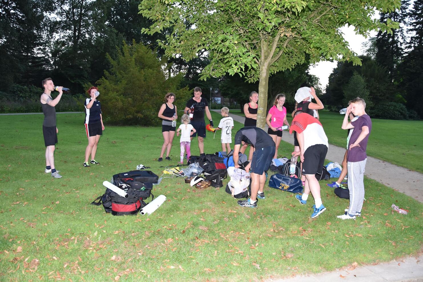 Gruppe von Sportlern in Laufkleidung versammelt unter einem Baum, mit Rucks&auml;cken und Ausr&uuml;stung auf einer Wiese.