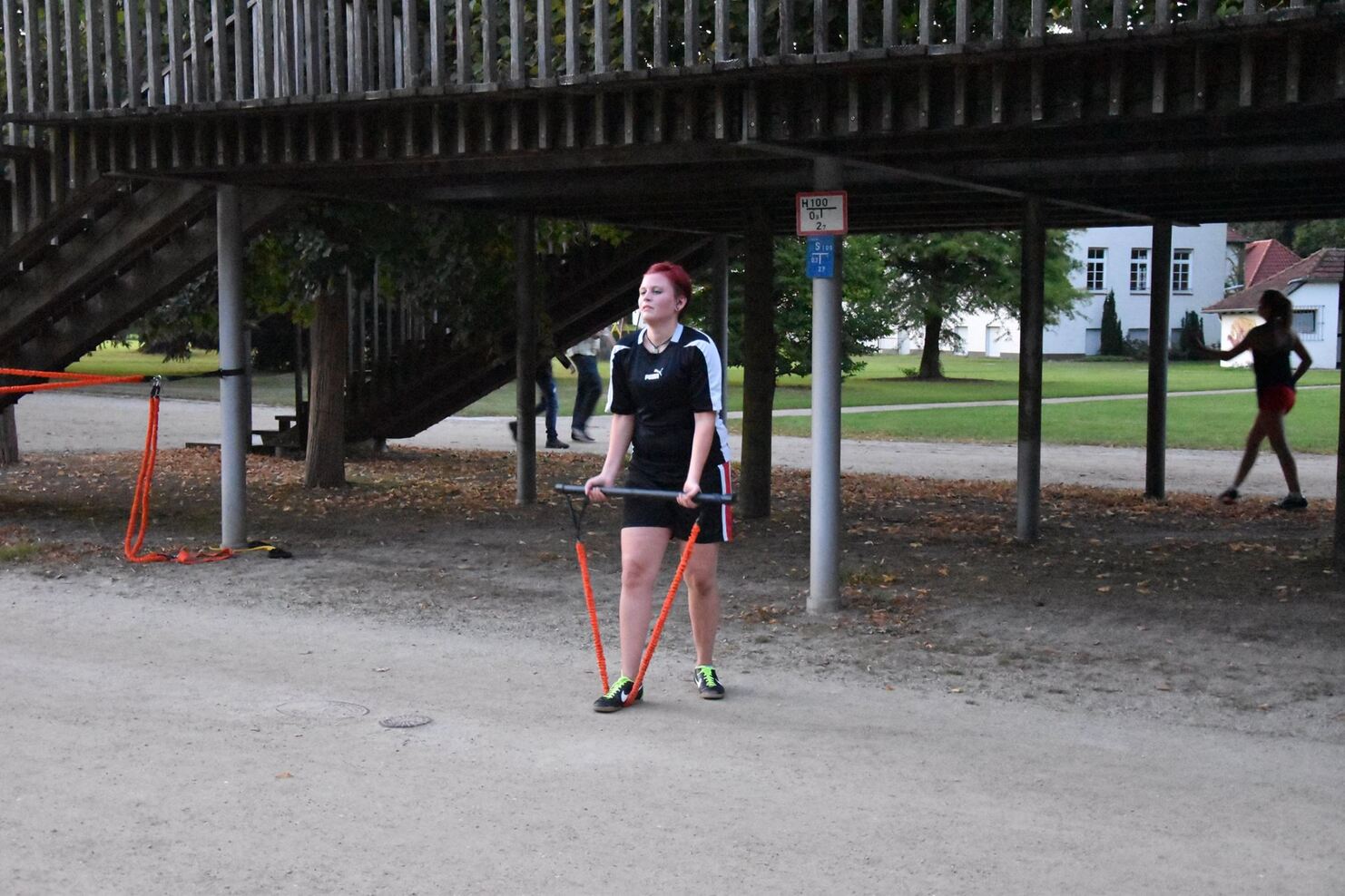 Frau mit roten Haaren in Sportkleidung h&auml;lt Trainingsb&auml;nder und steht auf einem Platz unter einem Spielplatz.