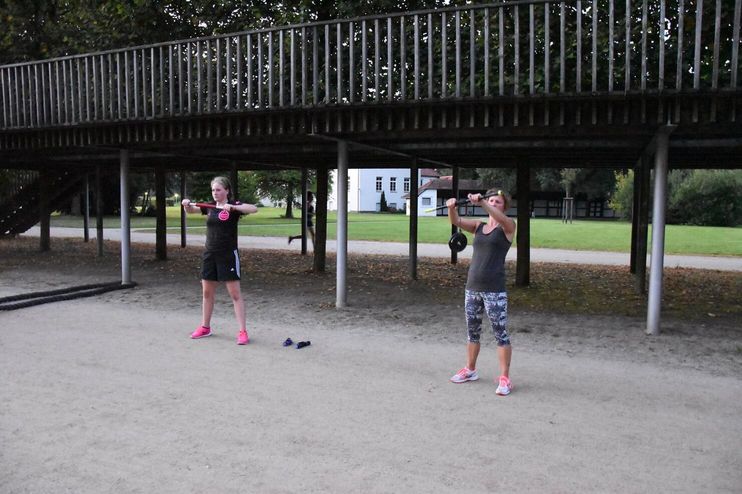 Zwei Frauen f&uuml;hren Kniebeugen mit Gewichten auf einem Sportplatz unter einem Holzger&uuml;st aus.