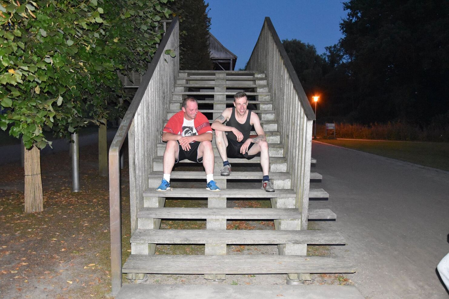 Zwei M&auml;nner sitzen auf den Stufen einer Holztreppe in der D&auml;mmerung, umgeben von B&auml;umen und einem beleuchteten Weg.
