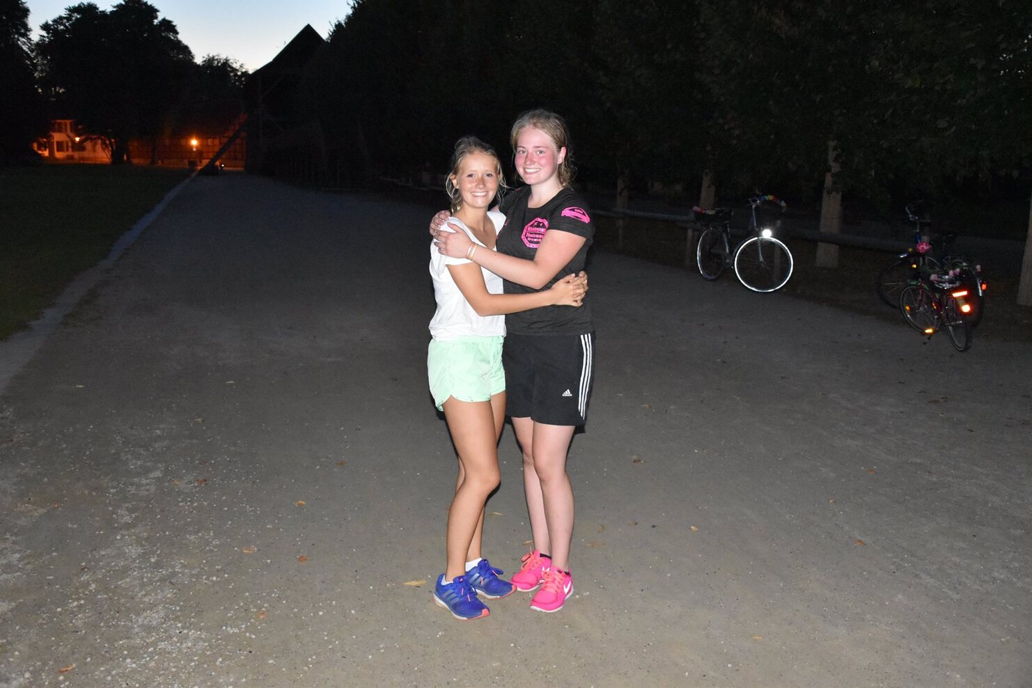 Zwei Jugendliche umarmen sich l&auml;chelnd auf einem beleuchteten Weg, im Hintergrund stehen Fahrr&auml;der.