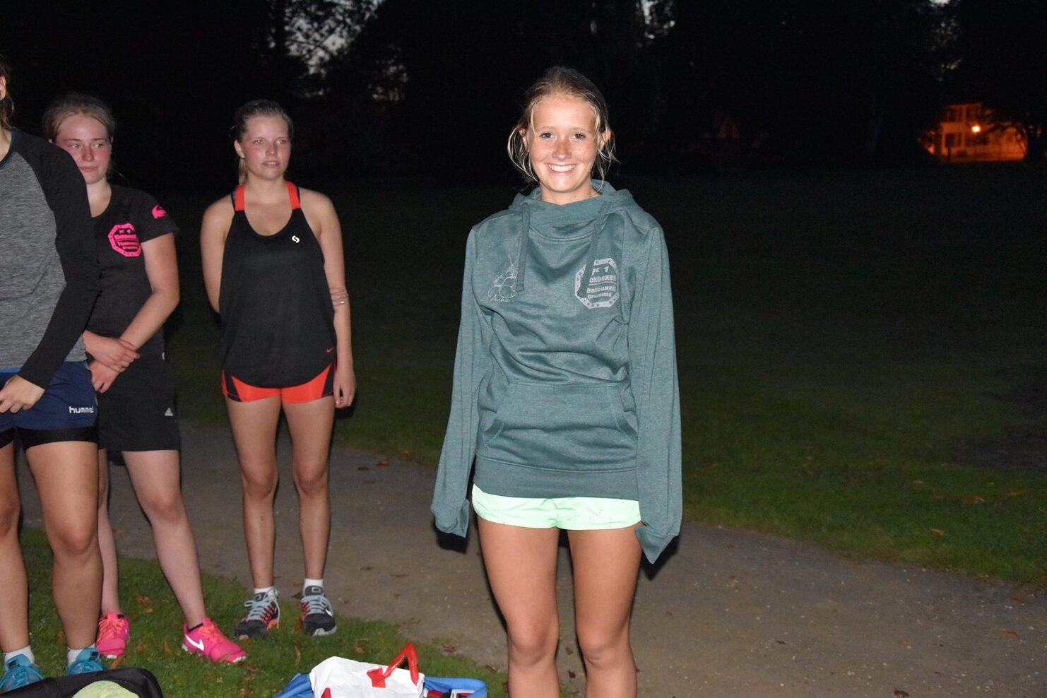 L&auml;chelnde Sportlerin in einer &uuml;bergro&szlig;en Sweatshirt steht im Vordergrund, w&auml;hrend andere Sportlerinnen im Hintergrund warten.