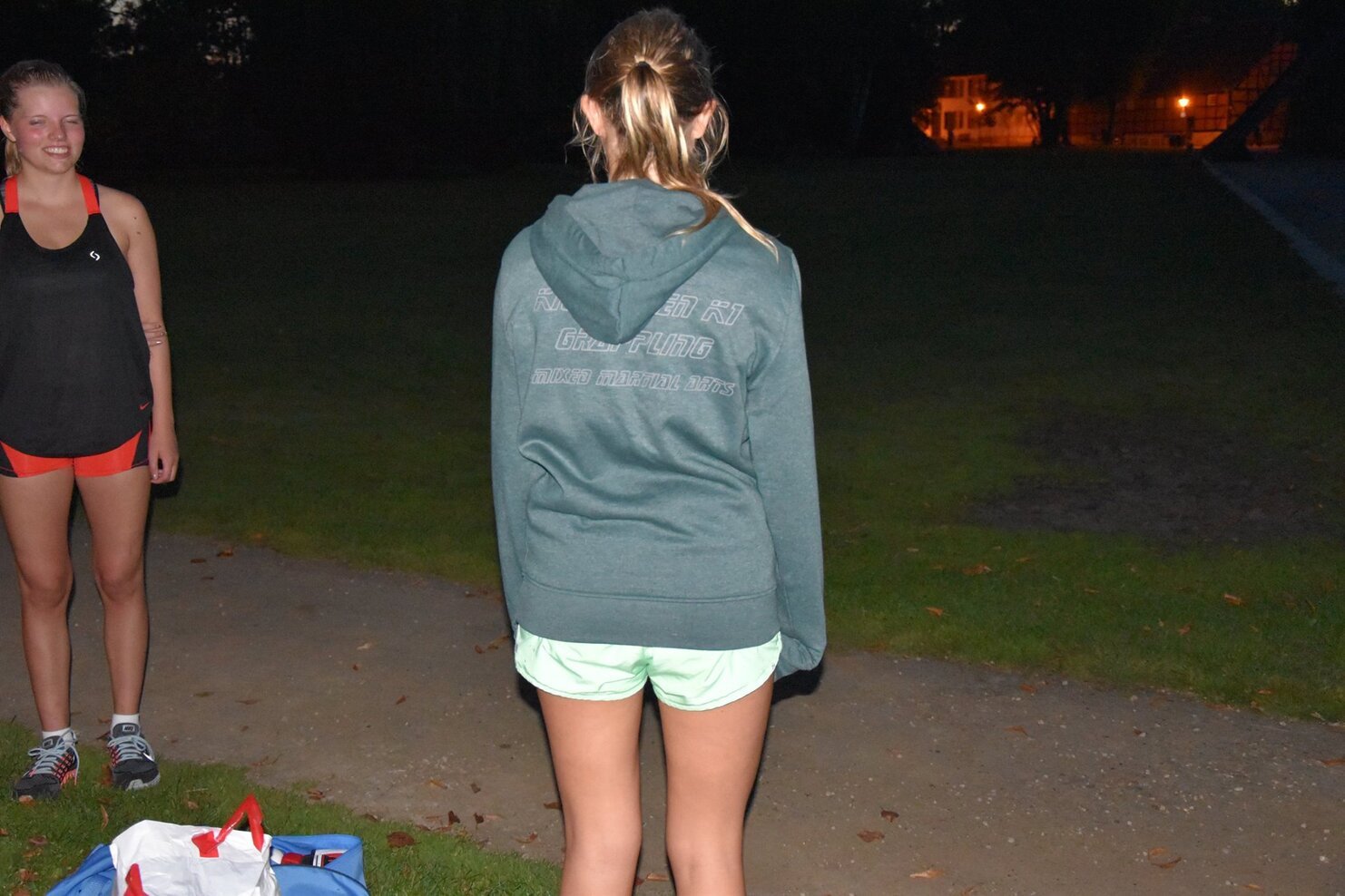 Zwei Sportlerinnen im Freien: Eine steht mit dem R&uuml;cken zur Kamera, die andere l&auml;chelt im Hintergrund. Dunkle Umgebung.