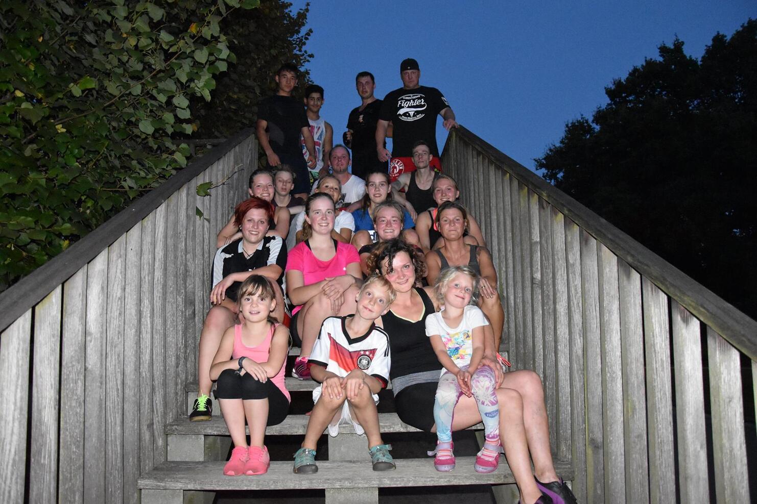 Gruppe von Kindern und Erwachsenen sitzt und steht auf einer Holztreppe in der D&auml;mmerung, umgeben von B&auml;umen.