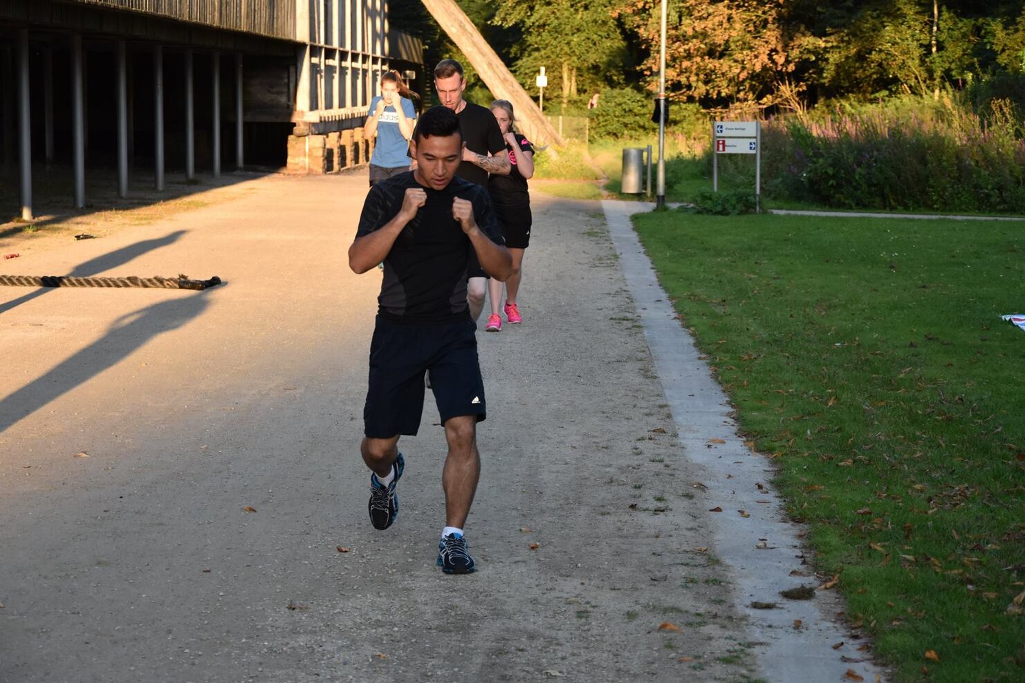 Mann in Sportkleidung l&auml;uft auf einem Weg, w&auml;hrend zwei weitere Personen im Hintergrund joggen. Tageslichtbeleuchtung.