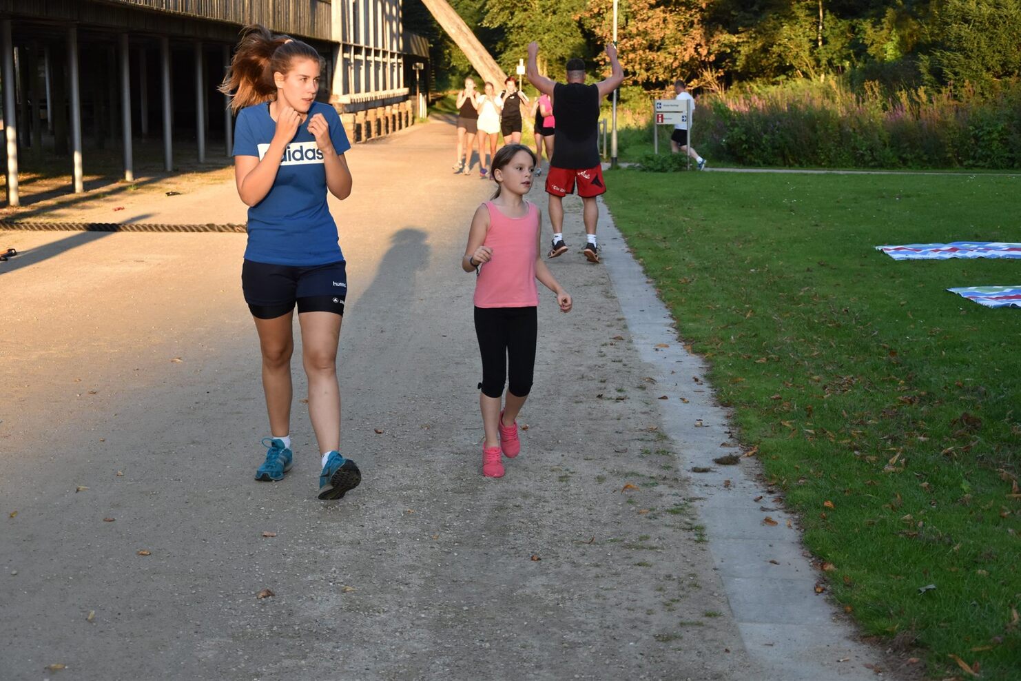 Zwei M&auml;dchen joggen auf einem Weg, umgeben von gr&uuml;nem Gras und B&auml;umen, w&auml;hrend andere im Hintergrund aktiv sind.