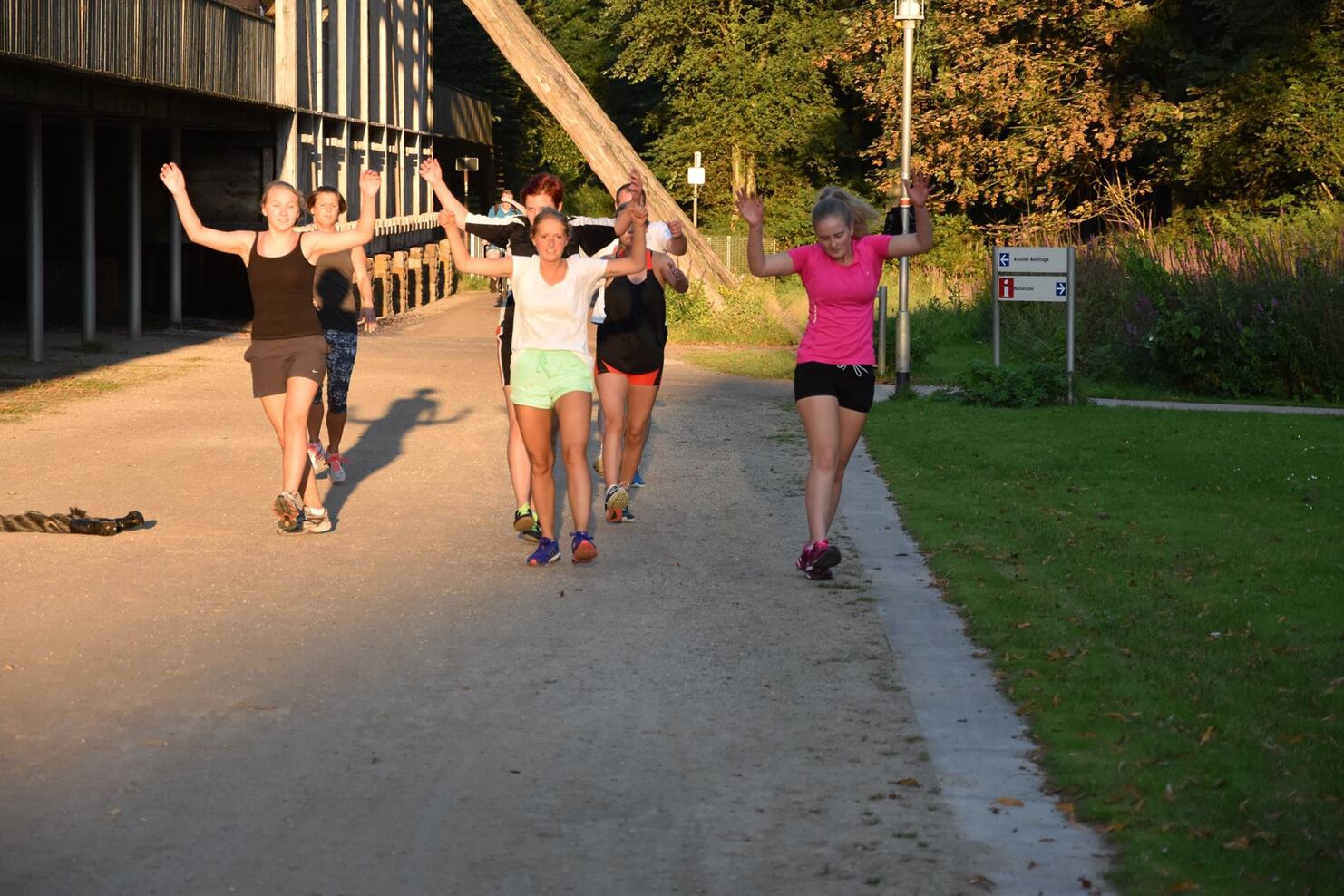 Gruppentraining mit sechs Frauen, die in Sportkleidung auf einem Weg joggen und fr&ouml;hlich die Arme heben.
