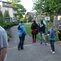 Gruppe von f&uuml;nf Personen auf einem Weg in einem Garten, umgeben von B&auml;umen und Wohngeb&auml;uden, mit Spielverbotsschild.