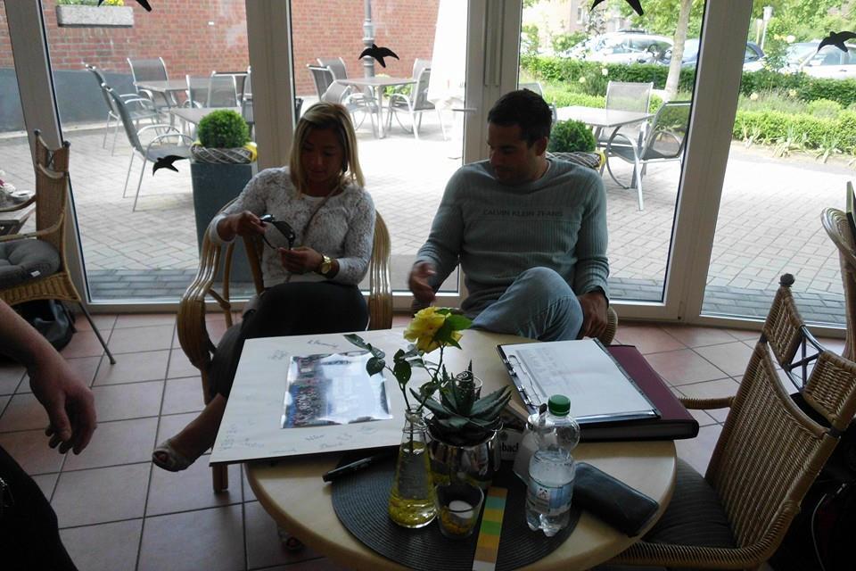 Zwei Personen sitzen an einem Tisch in einem hellen Raum, umgeben von Pflanzen und einem Blatt Papier.