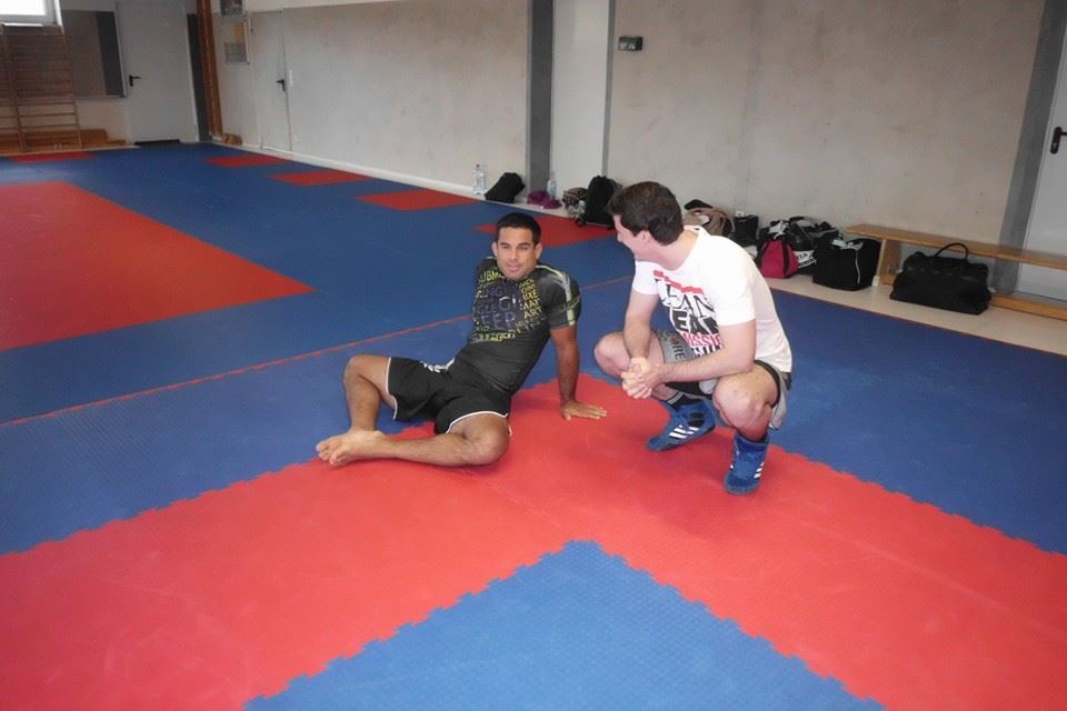 Zwei M&auml;nner im Trainingsraum: Einer sitzt auf dem Boden, der andere kniet daneben und spricht mit ihm.