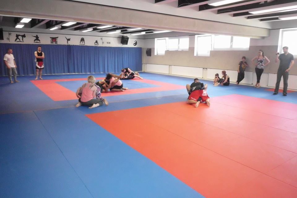 Trainierende k&auml;mpfen auf einer blauen und roten Matte in einem Sportraum, Zuschauer am Rand beobachten das Geschehen.