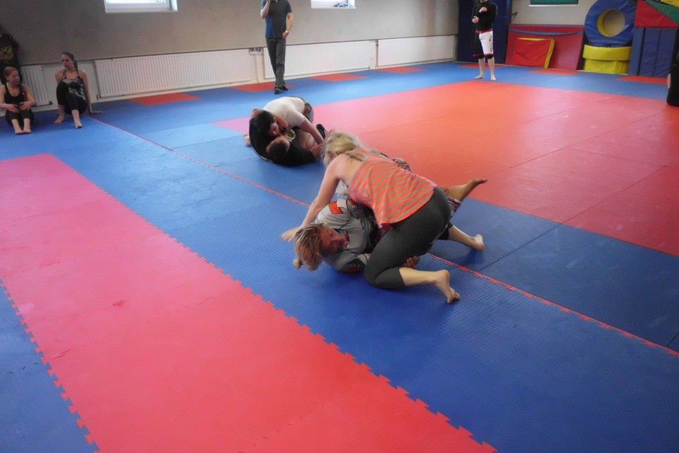 Zwei Personen k&auml;mpfen auf einer Kampfsportmatte, w&auml;hrend Zuschauer im Hintergrund sitzen und zuschauen.