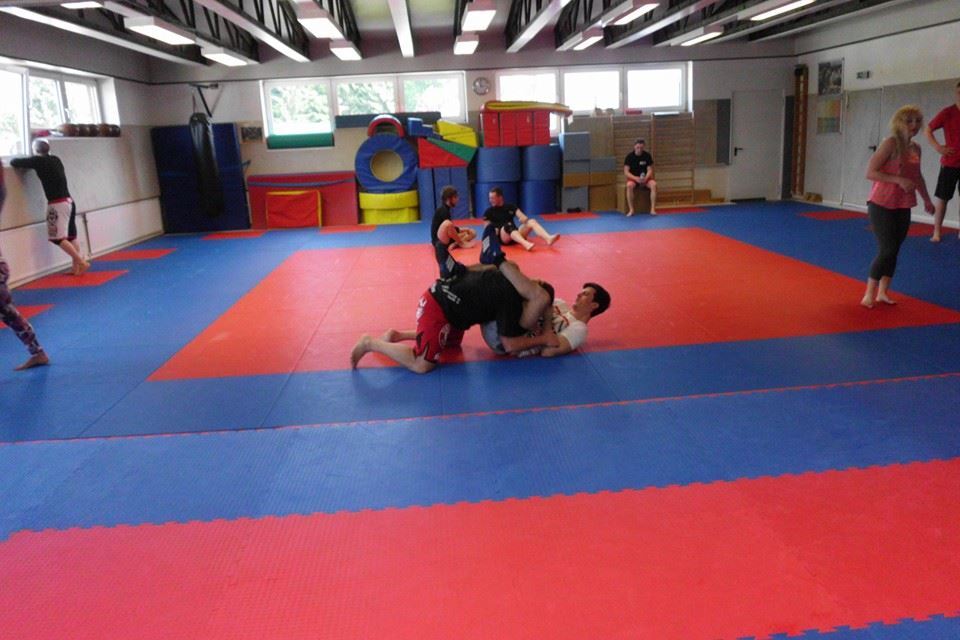 Trainingseinheit in einem Kampfsportraum mit mehreren Personen auf blauen und roten Matten, eine Gruppe &uuml;bt Bodentechniken.