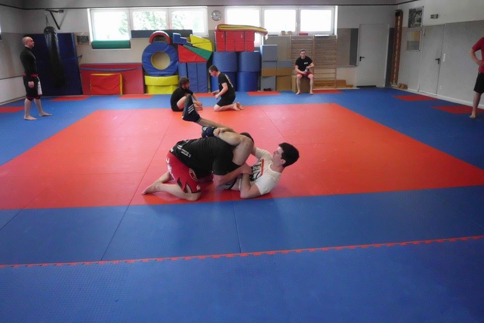Trainingsszene in einem Raum mit mehreren K&auml;mpfern, die auf einer blauen und roten Matte Grappling praktizieren.