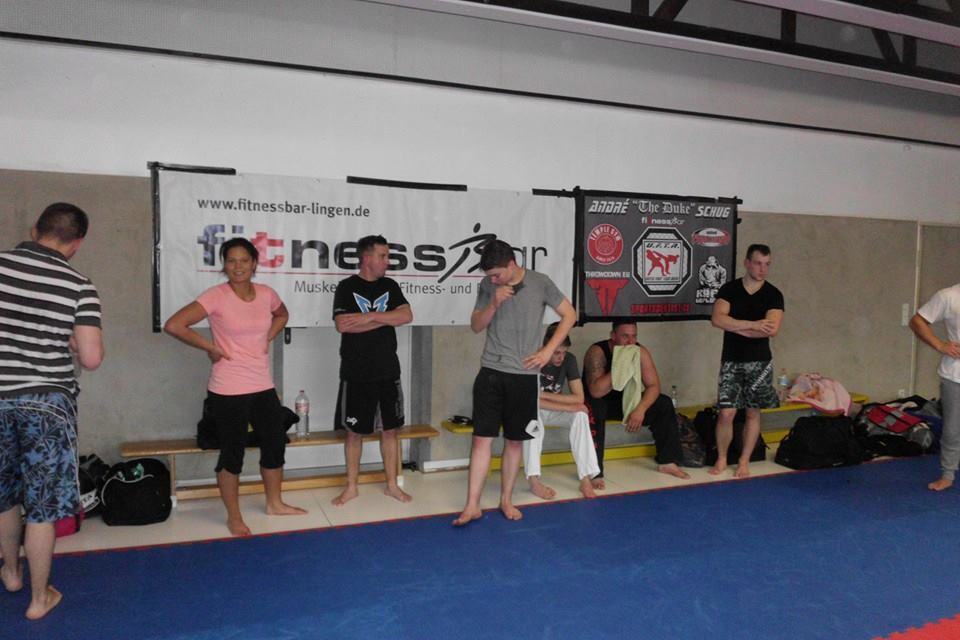 Gruppierung von Sportlern in Trainingskleidung, die an einer Wand mit Werbebannern stehen. Mattenboden sichtbar.