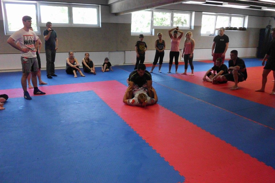 Gruppentraining in einer Kampfsporthalle mit mehreren Personen auf Matten, w&auml;hrend zwei Personen eine Technik durchf&uuml;hren.