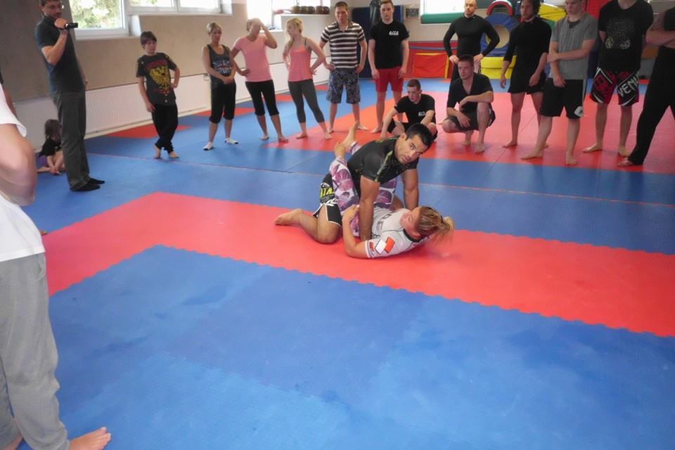 Training in einem Kampfsportraum: Ein Trainer zeigt eine Technik an einer Sportlerin, w&auml;hrend eine Gruppe zuschaut.