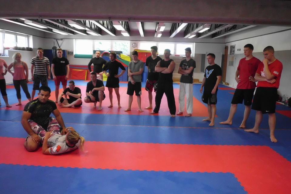 Zwei K&auml;mpfer demonstrieren eine Technik auf einer Trainingsmatte, w&auml;hrend eine Gruppe Zuschauer aufmerksam zusieht.