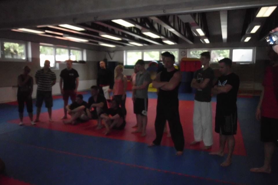 Gruppe von Personen in einem Kampfsportraum, die auf einem roten und blauen Mattenboden stehen und zuschauen.