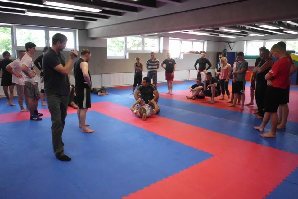 Gruppe von Menschen beobachtet eine Kampfsport-Demonstration auf einer blauen und roten Mattenfl&auml;che in einem Trainingsraum.