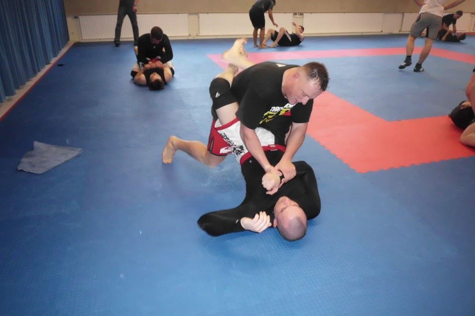 Zwei K&auml;mpfer im Bodenkampf auf einer blauen Matte, einer kontrolliert den anderen im Grappling-Stil.