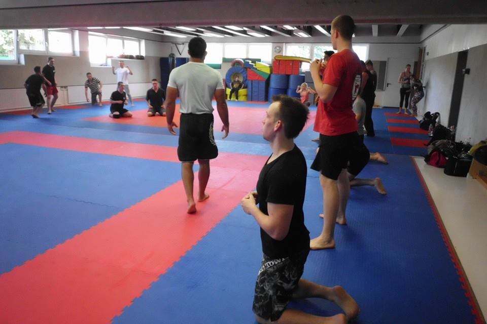Gruppentraining in einem Kampfsportraum; Teilnehmer &uuml;ben auf blauen und roten Matten, einige auf den Knien, andere stehen.