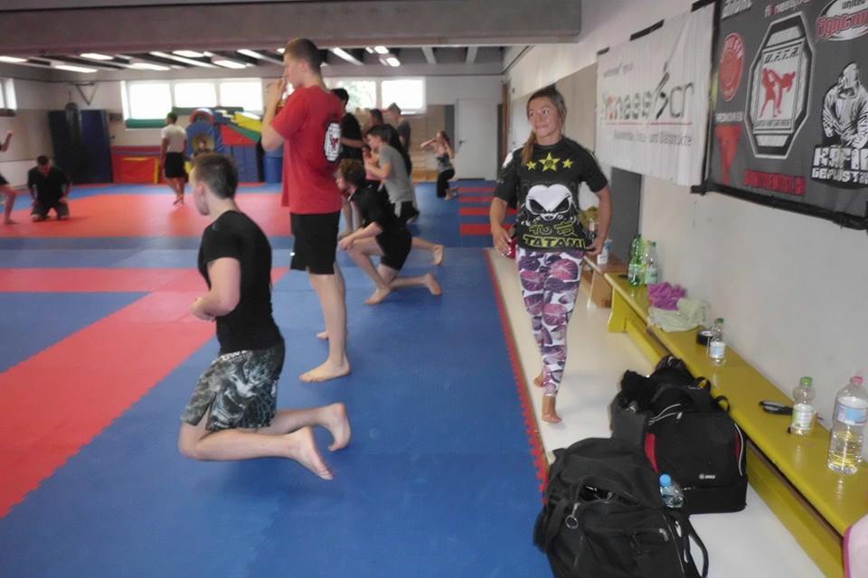 Sportlicher Trainingsraum mit mehreren Personen beim Training; eine Frau steht l&auml;chelnd im Vordergrund.