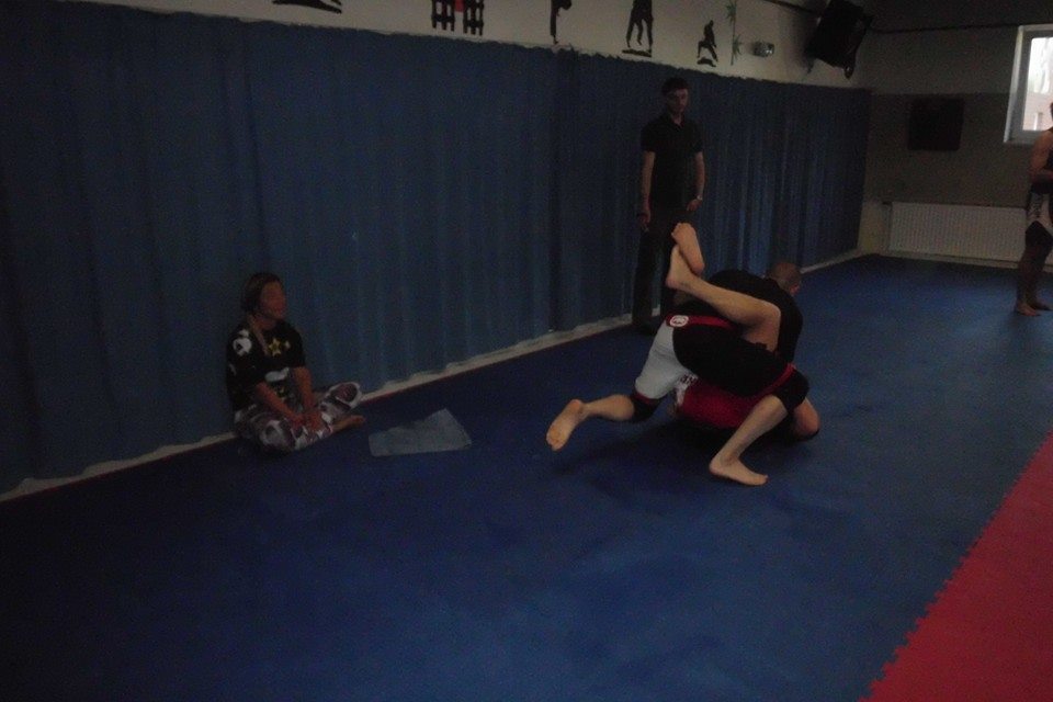 Zwei K&auml;mpfer in Sportkleidung k&auml;mpfen auf einer blauen Matratze, w&auml;hrend eine Person im Hintergrund zuschaut.