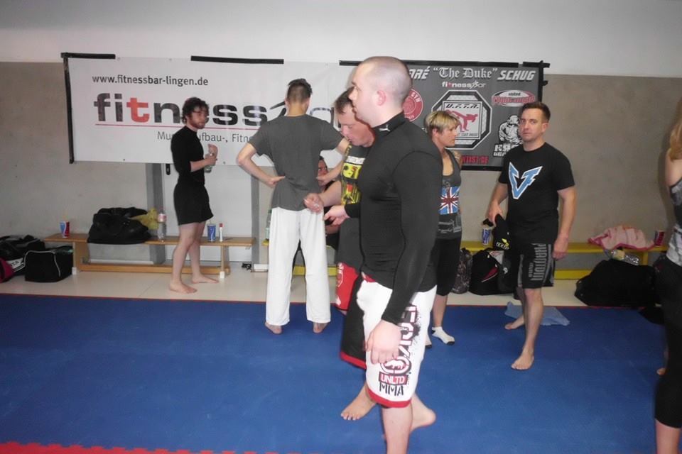 Gruppe von Kampfsportlern im Training, in sportlicher Kleidung, vor einer Wand mit Fitnessbanner in einem &Uuml;bungsraum.