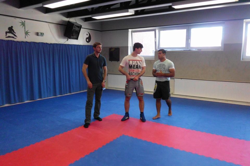 Drei M&auml;nner stehen in einem Trainingsraum mit blauen und roten Matten und diskutieren miteinander.