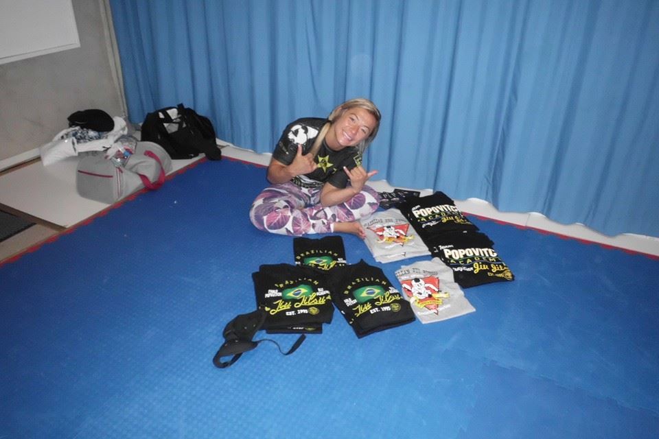 Frau im Sportoutfit sitzt auf einer Matte und zeigt das Peace-Zeichen neben mehreren T-Shirts auf dem Boden.