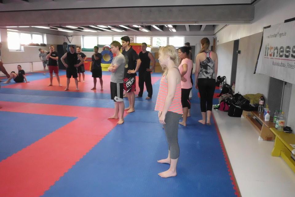 Gruppe junger Menschen in Trainingskleidung steht auf einer gepolsterten Matte in einem Sportraum, Diskussion im Gange.