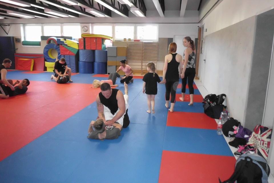 Gruppentraining auf einer Matte, Menschen in Schwarz und Grau &uuml;ben Techniken, Kinder spielen im Hintergrund.