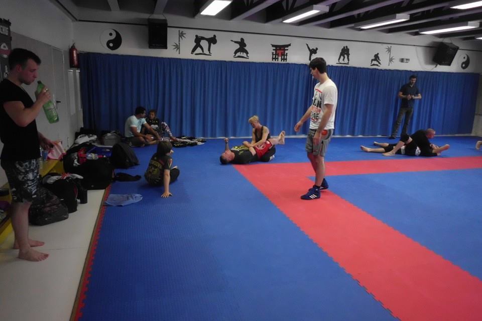 Trainierende in einem Kampfsportraum mit blauen Matten, w&auml;hrend einige auf dem Boden liegen und andere stehen.