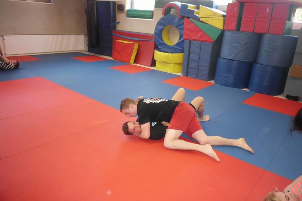 Zwei Jugendliche &uuml;ben auf einer Matte, einer h&auml;lt den anderen in einer Wrestling-Position. Bunte &Uuml;bungsger&auml;te im Hintergrund.