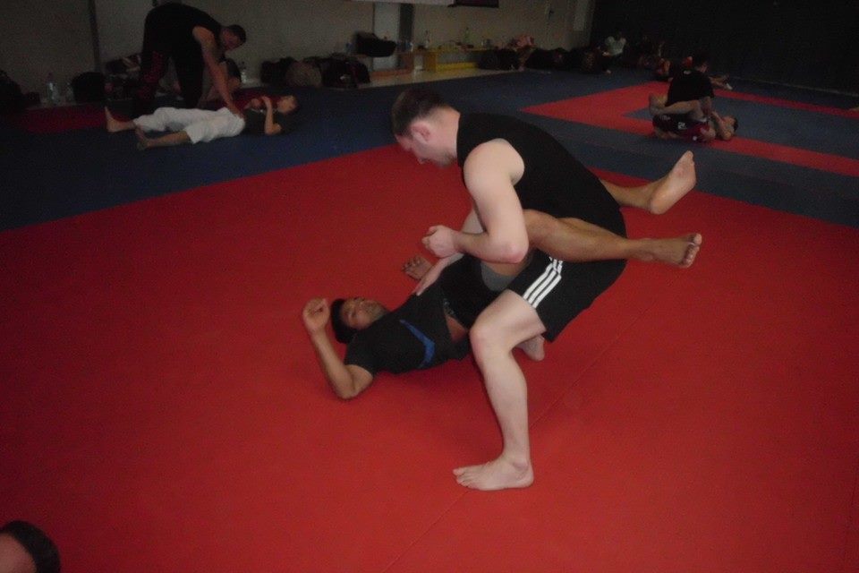 Zwei Sportler trainieren auf einer roten Trainingsmatte, einer f&uuml;hrt eine Haltegriff-&Uuml;bung aus.